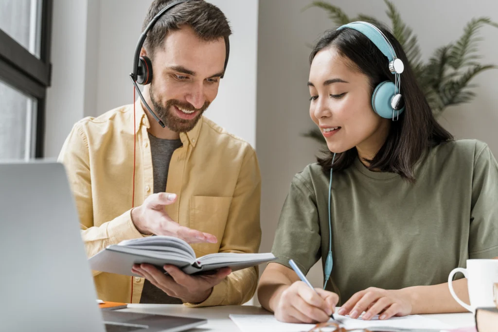 Alt text: A man and a woman wearing headsets are studying together at a desk. The man, holding an open notebook, gestures as he explains something, while the woman takes notes with a pen and smiles. A laptop, mug, and plant are visible in the background, suggesting an online learning or virtual meeting environment.